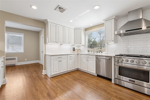 kitchen featuring radiator, stainless steel appliances, wall chimney range hood, visible vents, and light wood-style floors
