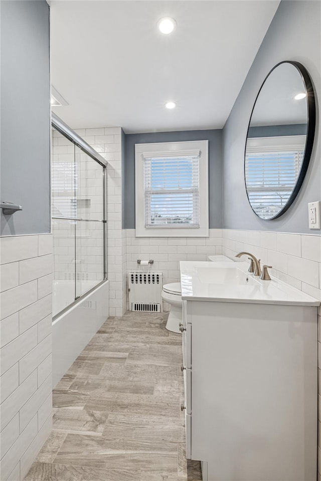 full bathroom featuring tile walls, toilet, vanity, radiator heating unit, and shower / bath combination with glass door
