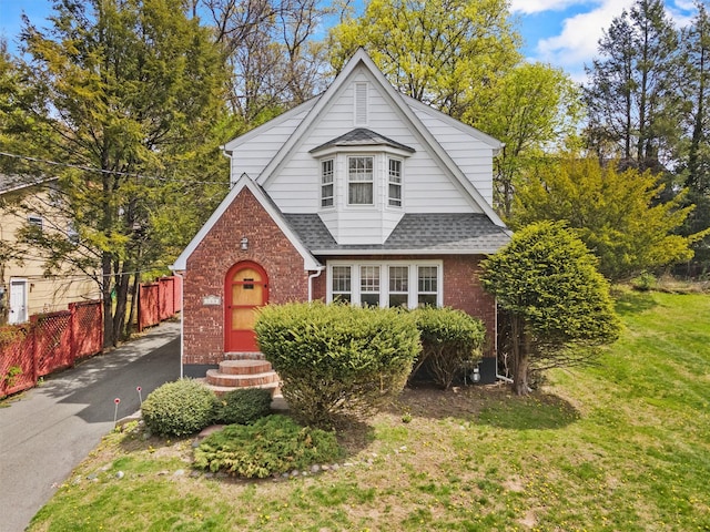 english style home with brick siding, fence, a front lawn, and roof with shingles