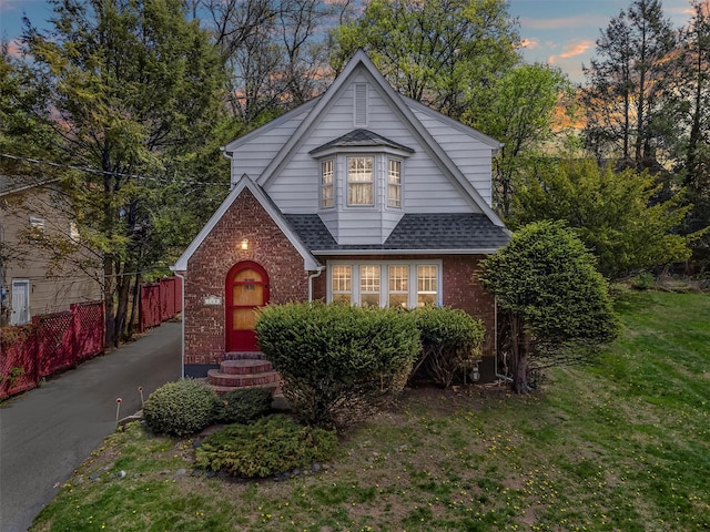 tudor home with a front yard, brick siding, roof with shingles, entry steps, and fence
