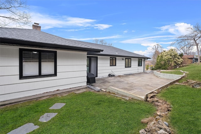 back of property with a lawn, roof with shingles, a chimney, and a patio