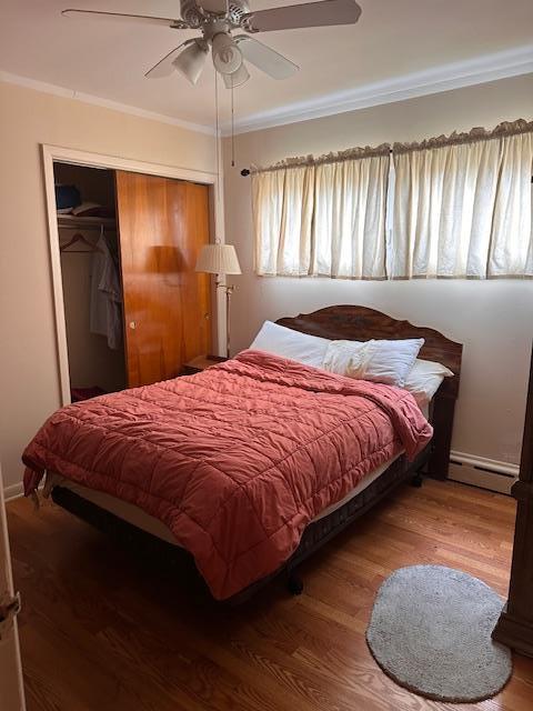 bedroom with wood finished floors, a ceiling fan, a closet, crown molding, and baseboard heating