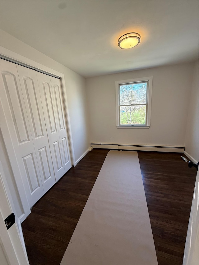 unfurnished bedroom featuring a baseboard heating unit, baseboards, a closet, and dark wood-style floors
