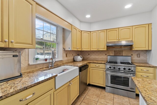 kitchen with appliances with stainless steel finishes, under cabinet range hood, a sink, light stone counters, and backsplash