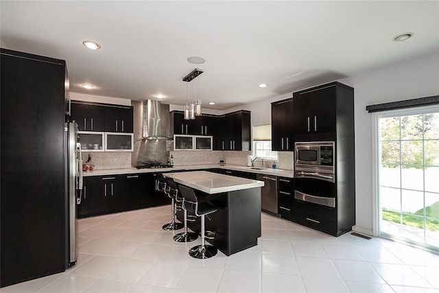 kitchen with wall chimney exhaust hood, dark cabinetry, light countertops, a kitchen breakfast bar, and recessed lighting