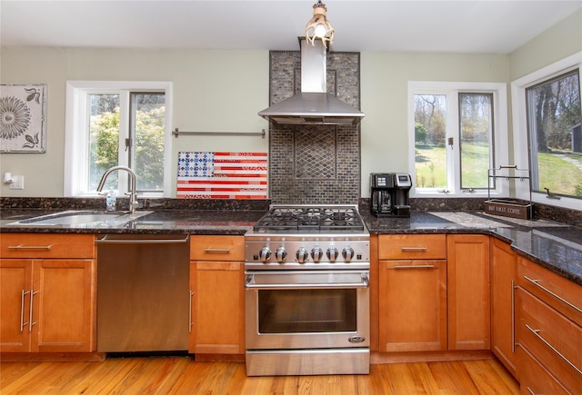kitchen with brown cabinetry, appliances with stainless steel finishes, a sink, dark stone countertops, and wall chimney exhaust hood