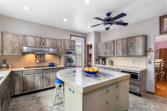 kitchen featuring under cabinet range hood, black electric stovetop, tile counters, oven, and a ceiling fan