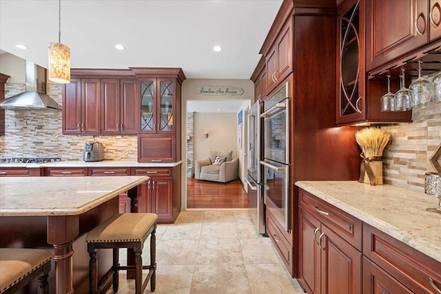 kitchen featuring stainless steel appliances, reddish brown cabinets, pendant lighting, wall chimney range hood, and glass insert cabinets