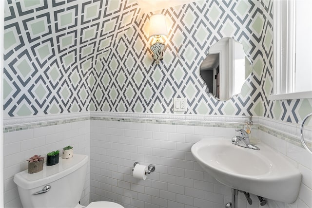 half bathroom featuring a sink, tile walls, toilet, and wallpapered walls
