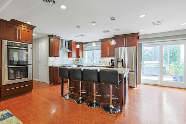 kitchen featuring light wood-style flooring, appliances with stainless steel finishes, a kitchen island, wall chimney range hood, and visible vents