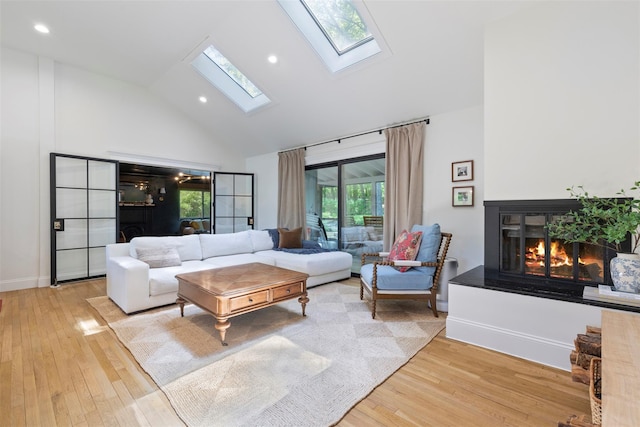 living area with high vaulted ceiling, a skylight, light wood finished floors, a glass covered fireplace, and baseboards