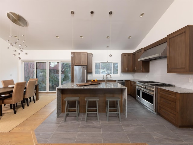 kitchen featuring backsplash, a center island with sink, wall chimney exhaust hood, appliances with stainless steel finishes, and a sink