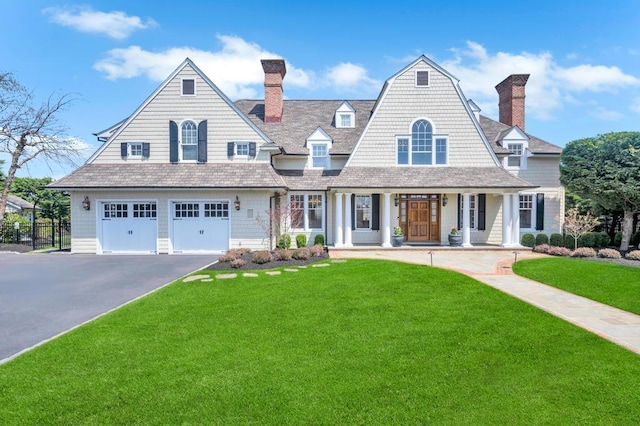 shingle-style home featuring a garage, aphalt driveway, covered porch, a front yard, and a chimney