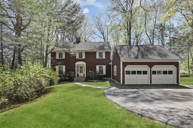colonial-style house featuring roof with shingles, an attached garage, a front lawn, a chimney, and driveway