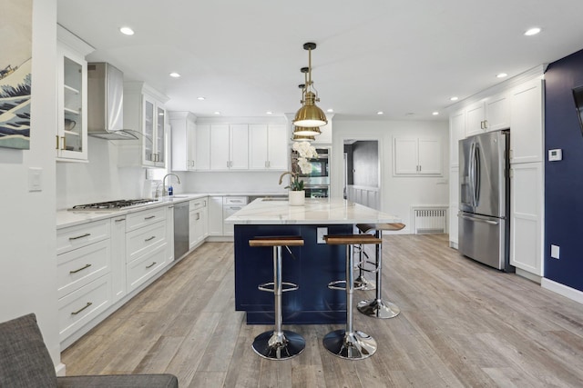 kitchen with wall chimney range hood, white cabinets, light wood-type flooring, stainless steel appliances, and radiator