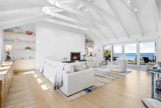 living room with beam ceiling, a lit fireplace, high vaulted ceiling, a water view, and light wood-style flooring