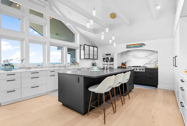 kitchen with light wood-style floors, an island with sink, tasteful backsplash, beamed ceiling, and modern cabinets