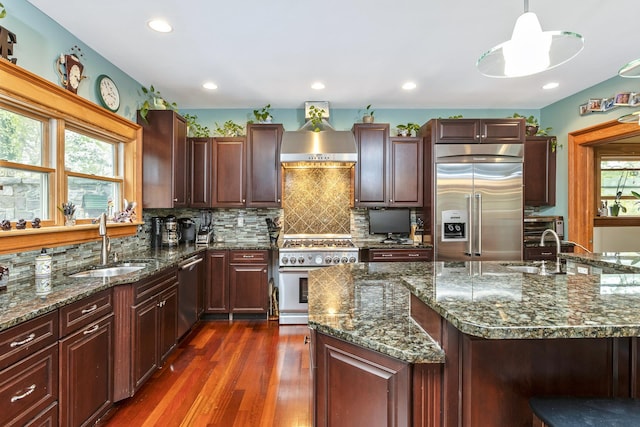 kitchen with wall chimney range hood, backsplash, a sink, premium appliances, and dark wood-type flooring