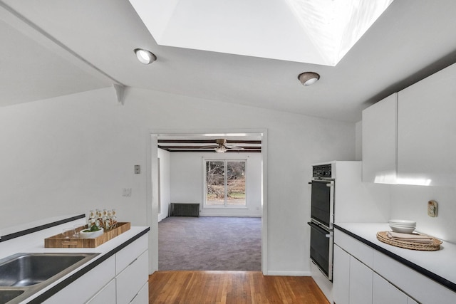 kitchen featuring multiple ovens, white cabinets, light wood finished floors, baseboards, and lofted ceiling with skylight