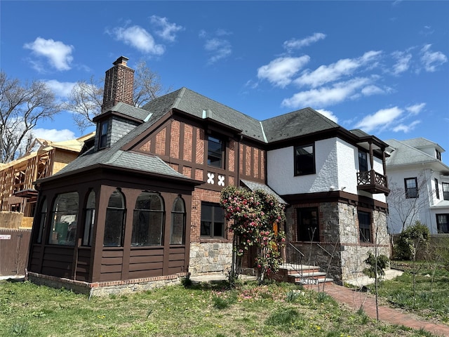 back of house featuring roof with shingles, a balcony, stone siding, and a chimney