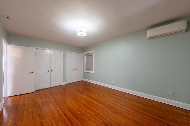 unfurnished bedroom with two closets, wood finished floors, baseboards, and an AC wall unit