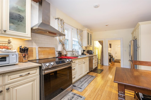kitchen featuring wall chimney range hood, cream cabinetry, a sink, stainless steel appliances, and backsplash