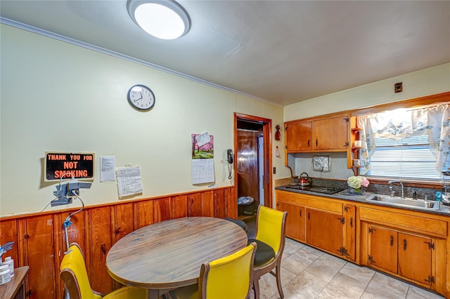 kitchen with black electric cooktop, a wainscoted wall, a sink, light tile patterned flooring, and brown cabinetry