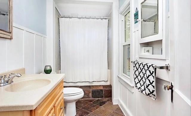 bathroom with a decorative wall, vanity, a wainscoted wall, toilet, and shower / bath combo