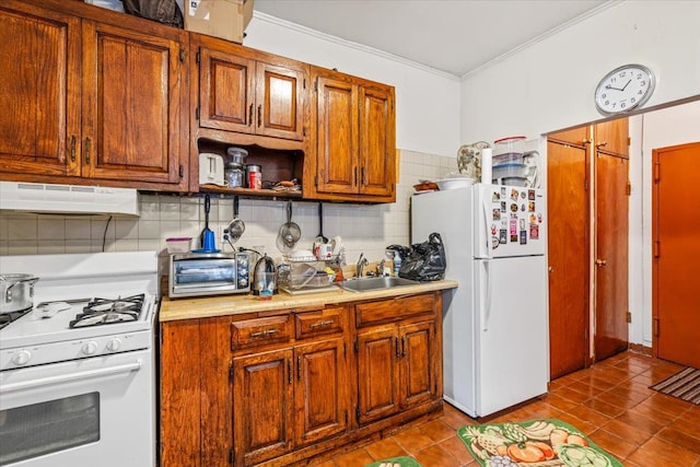 kitchen with a sink, under cabinet range hood, ornamental molding, dark tile patterned flooring, and white appliances