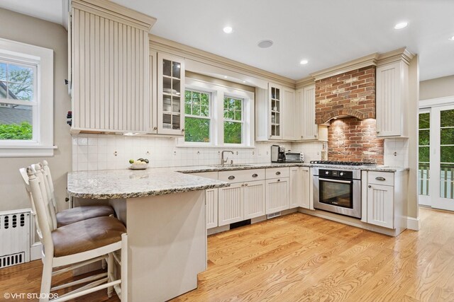 kitchen featuring appliances with stainless steel finishes, a peninsula, light wood-type flooring, glass insert cabinets, and decorative backsplash