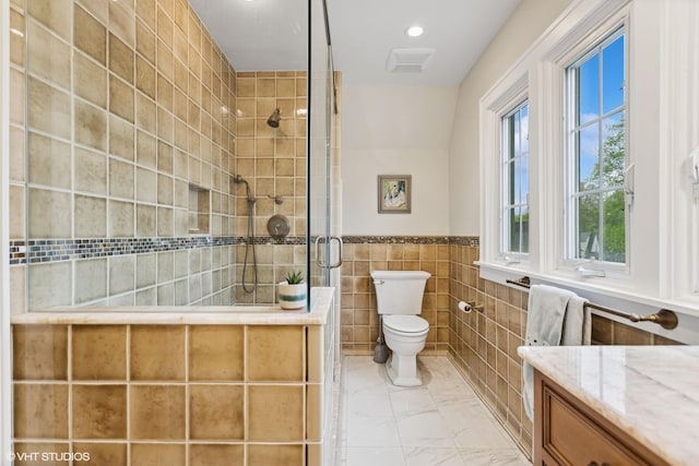 bathroom featuring tiled shower, tile walls, marble finish floors, toilet, and wainscoting