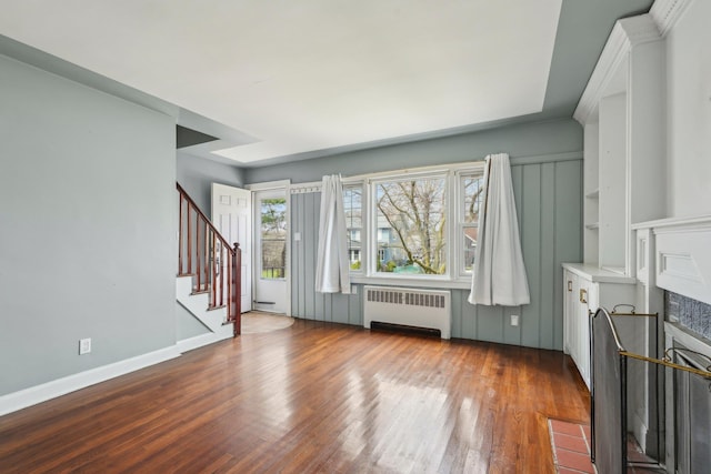 unfurnished living room with baseboards, stairs, a fireplace with flush hearth, radiator, and wood finished floors