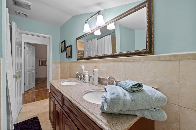 bathroom with tile walls, a wainscoted wall, tile patterned flooring, double vanity, and vaulted ceiling