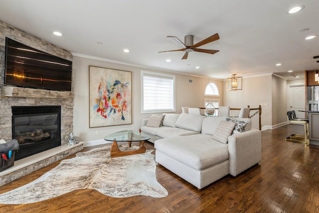 living room with ceiling fan, dark wood finished floors, ornamental molding, a stone fireplace, and recessed lighting