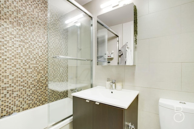 bathroom with bath / shower combo with glass door, vanity, toilet, and tile walls