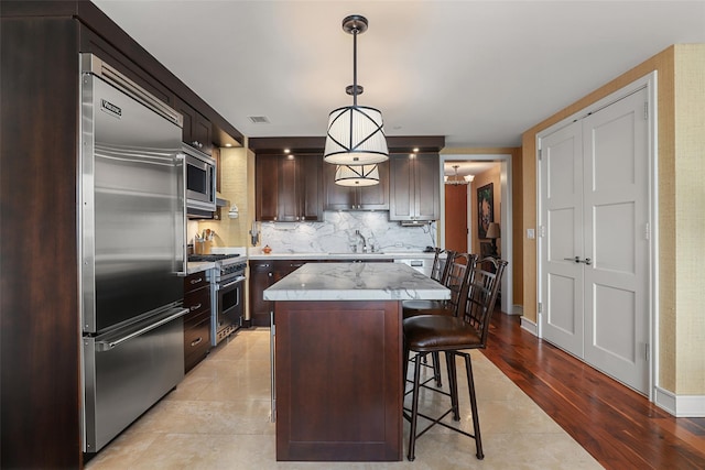 kitchen featuring a sink, built in appliances, dark brown cabinetry, backsplash, and a center island