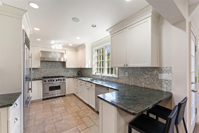 kitchen featuring a peninsula, a sink, high end appliances, backsplash, and wall chimney range hood