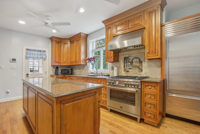 kitchen featuring brown cabinetry, a ceiling fan, high end appliances, a sink, and wall chimney exhaust hood