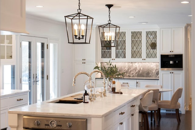 kitchen with light stone counters, a center island with sink, white cabinetry, backsplash, and hanging light fixtures