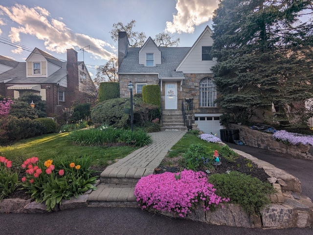 english style home with driveway, stone siding, a chimney, and an attached garage