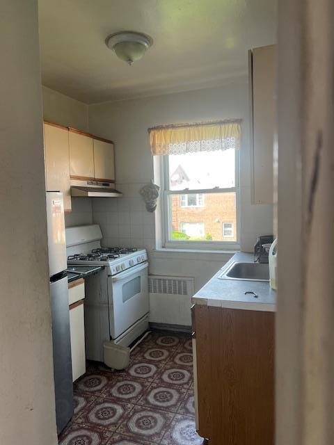 20-25 Parsons Blvd, Whitestone, NY 11357 - kitchen with white gas stove, radiator, freestanding refrigerator, under cabinet range hood, and light countertops