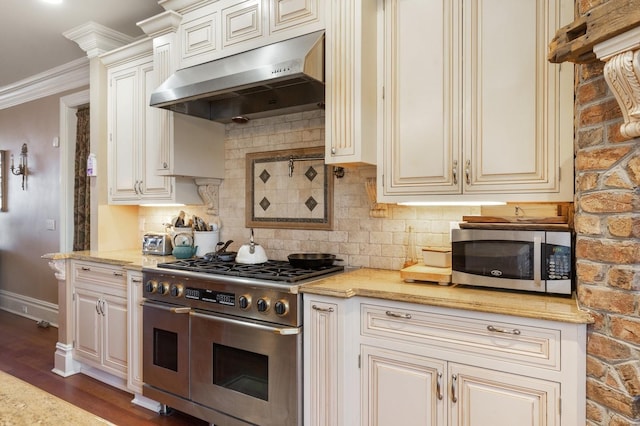 kitchen with cream cabinetry, under cabinet range hood, dark wood finished floors, stainless steel appliances, and decorative backsplash