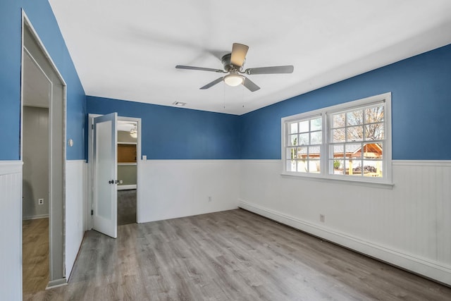 unfurnished bedroom featuring baseboard heating, wood finished floors, a wainscoted wall, and a ceiling fan