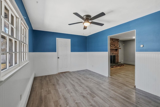 empty room with a fireplace, wood finished floors, a ceiling fan, and wainscoting