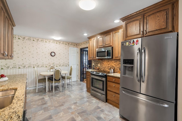 kitchen featuring light stone counters, stainless steel appliances, wallpapered walls, recessed lighting, and wainscoting