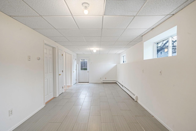 below grade area with a baseboard radiator, a drop ceiling, and baseboards