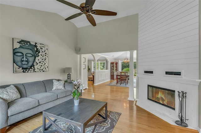 living area featuring light wood finished floors, a ceiling fan, arched walkways, a glass covered fireplace, and high vaulted ceiling