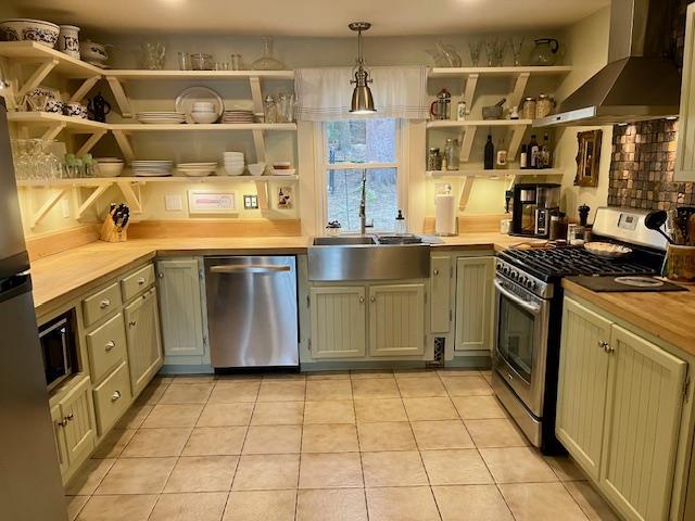 kitchen featuring stainless steel appliances, open shelves, wall chimney range hood, light tile patterned floors, and a sink