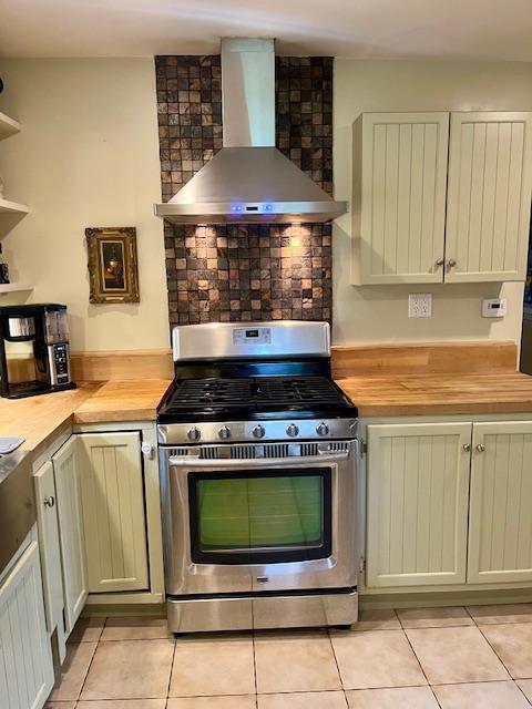 kitchen featuring open shelves, stainless steel gas range, light tile patterned floors, butcher block counters, and wall chimney exhaust hood
