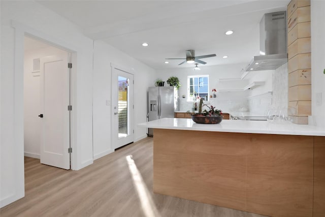 kitchen with wall chimney range hood, decorative backsplash, light wood-style floors, a peninsula, and stainless steel refrigerator with ice dispenser